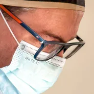 Dr. Lucas Schmidt de perfil, de óculos e máscara cirúrgica, olhando para baixo